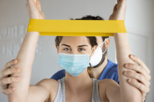 Young woman performing rehabilitative shoulder exercises using elastic band, with physiotherapist in a clinic, both wearing a face mask