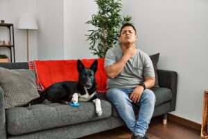 Young man sitting on sofa with dog and touching his painful throat