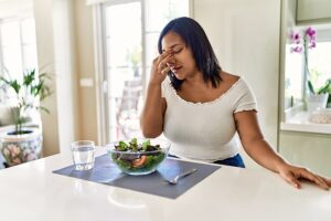 Woman sitting at table eating a salad and touching her forehead