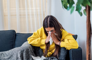 Young woman sick at home sitting on sofa under blanket and blowing her nose